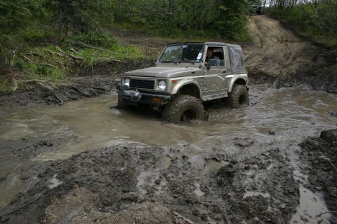 2003 Jeep TJ - An off-road vehicle navigates a challenging muddy trail, with another vehicle visible further up the incline.