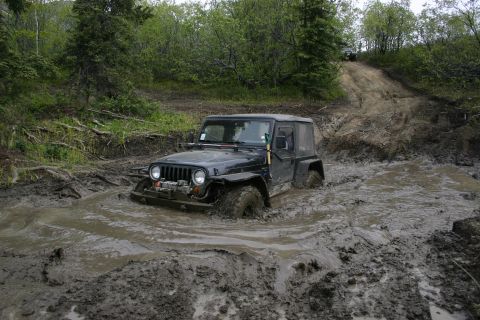 2003 Jeep TJ - A black 2003 Jeep TJ navigates a challenging muddy off-road course, with its tires submerged in thick mud.