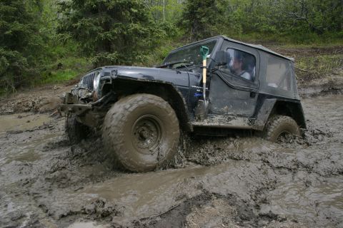 2003 Jeep TJ - A black Jeep Wrangler is deeply mired in a muddy trail, with its tires submerged and struggling for traction.