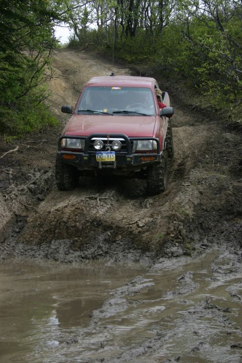 2003 Jeep TJ - A red pickup truck navigates a muddy trail with trees on either side.