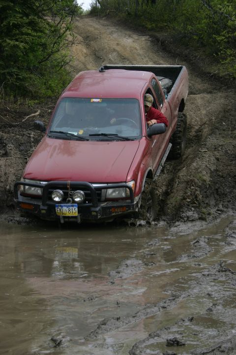 2003 Jeep TJ - A red pickup truck navigates a muddy off-road trail, its tires deep in the mire.