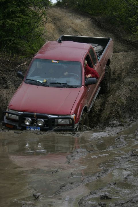 2003 Jeep TJ - A red pickup truck navigates a muddy trail, with its front tires deep in the mire.