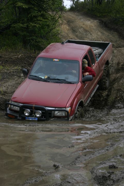 2003 Jeep TJ - This red Toyota Tacoma is stuck in the mud on a trail, with trees and a muddy incline in the background.