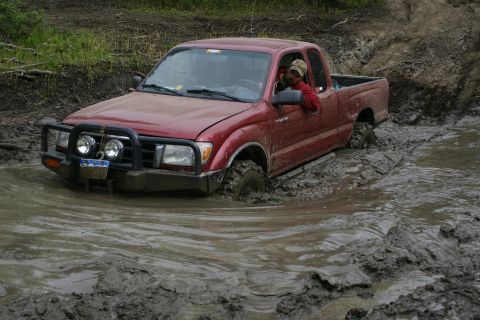 2003 Jeep TJ - A red pickup truck navigates a challenging muddy terrain, with the driver visible behind the wheel.