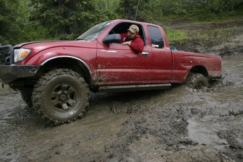 2003 Jeep TJ - A red pickup truck navigates a muddy off-road trail, its tires submerged in thick mud.