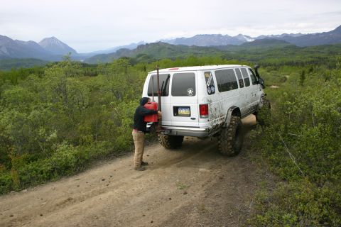 2003 Jeep TJ - A white van equipped for off-road travel is parked on a dirt path, with a person attending to a jerrycan mounted on its rear.