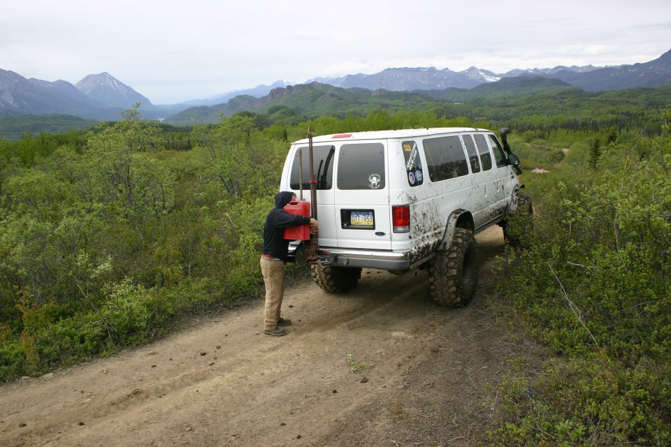 White van with off-road tires and jerrycan on rear, parked on a dirt road with mountains in background