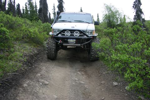 2003 Jeep TJ - A modified van with off-road tires navigates a rough dirt track surrounded by dense foliage and trees.