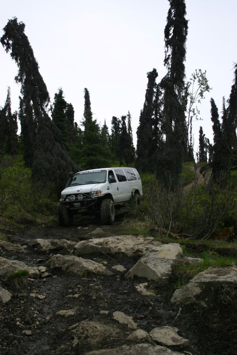 2003 Jeep TJ - A modified van navigates a challenging, rocky, and muddy off-road path through a forest.