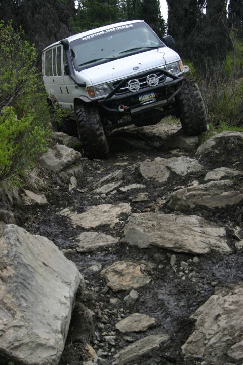 2003 Jeep TJ - This modified Ford van is tackling a challenging off-road course with large rocks and mud.