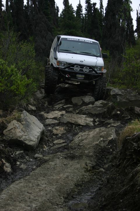 2003 Jeep TJ - A white off-road van tackles a challenging, rocky path in a wooded area.