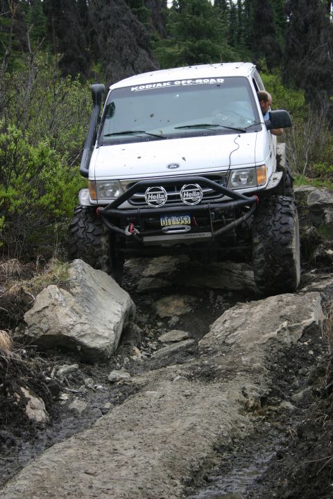 2003 Jeep TJ - A modified Ford van navigates a rocky, muddy off-road trail in a wooded area.