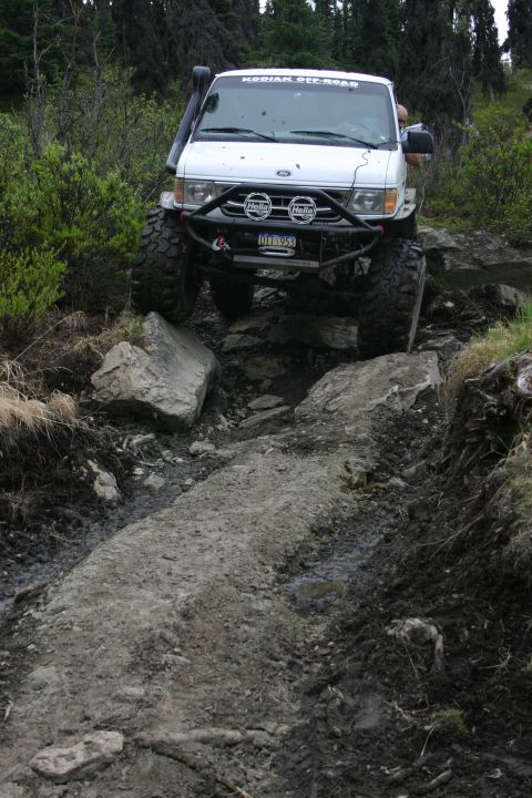 2003 Jeep TJ - A modified van tackles a challenging off-road path with large rocks and mud.