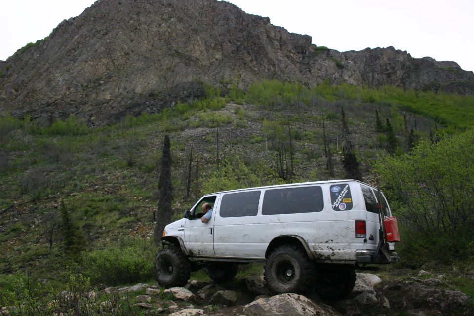 White van with big tires on a rocky trail, mountain in background. Has a red gas can on the back