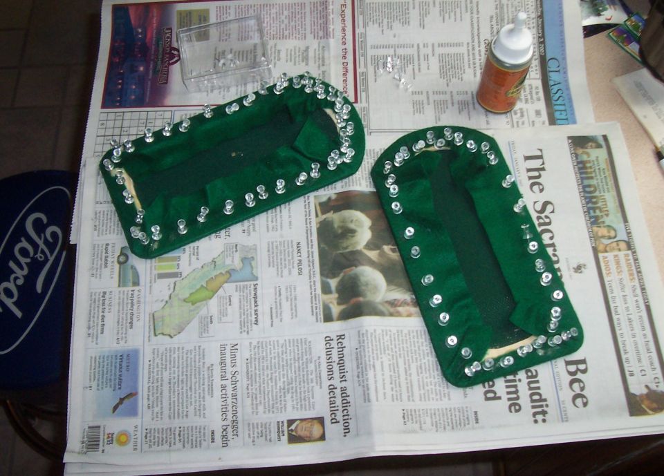 Two green fabric items with sequins on newspaper, a clear box of sequins, and a bottle of glue