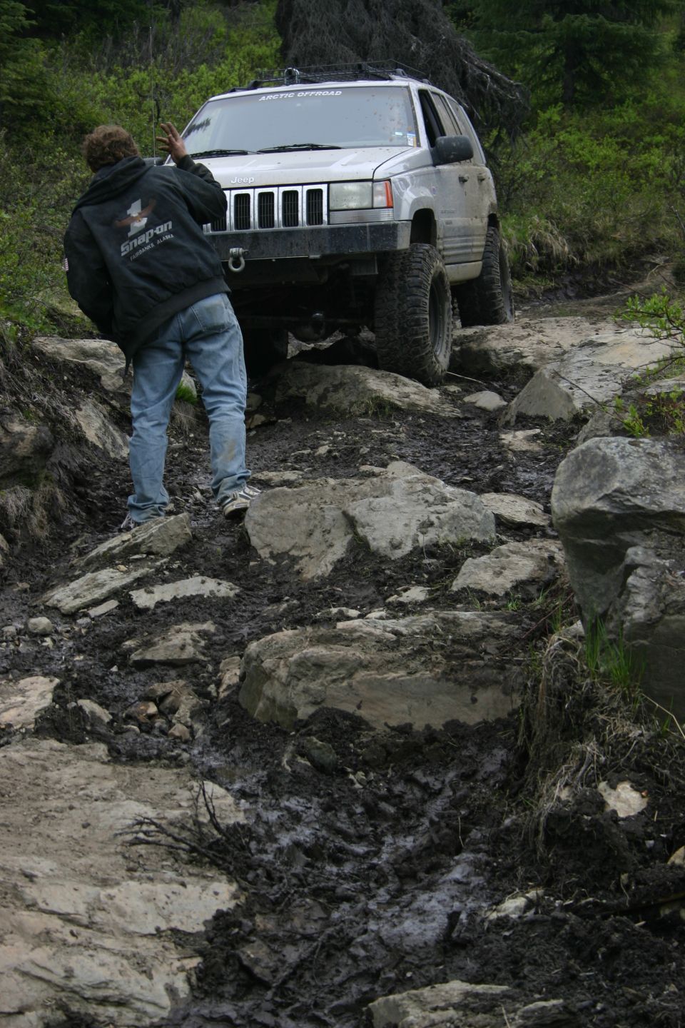 Silver Jeep Grand Cherokee on rocky, muddy trail. Person in Snap-On jacket guiding