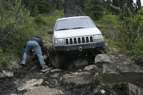 2003 Jeep TJ - A white Jeep Grand Cherokee maneuvers over rocks and mud on an off-road trail, with a person assisting near the front tire.