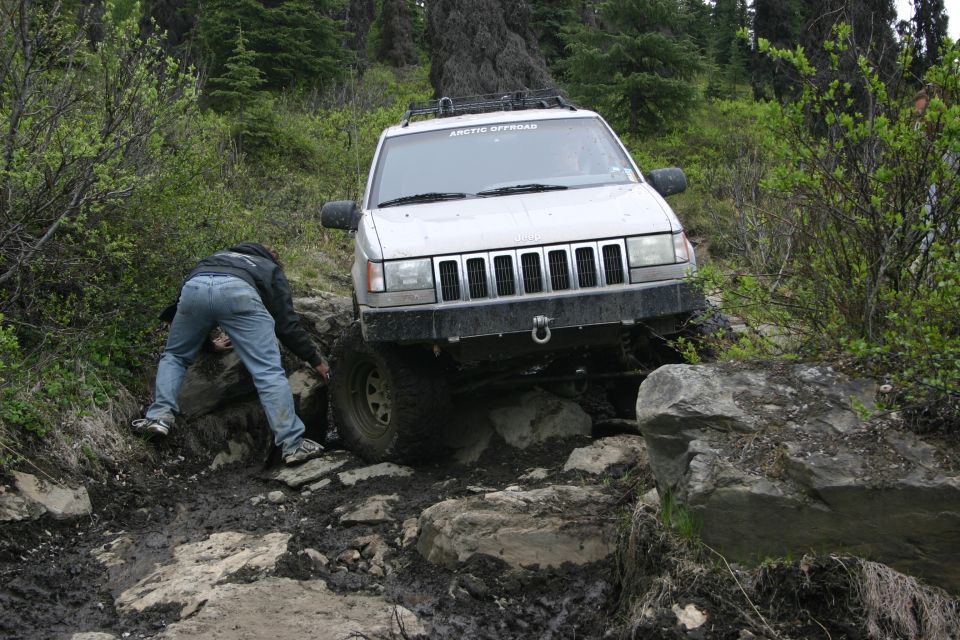 White Jeep Grand Cherokee navigating a rocky, muddy trail; "ARCTIC OFFROAD" sticker on windshield