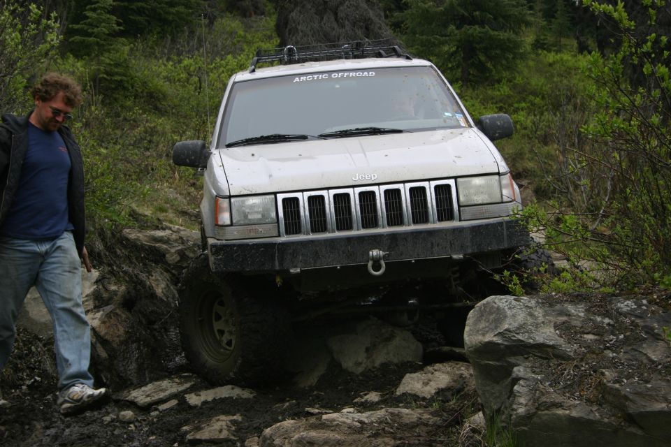 Silver Jeep Grand Cherokee navigating rocky terrain, with 'ARCTIC OFFROAD' sticker on windshield