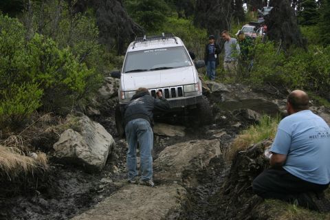 2003 Jeep TJ - A group of off-road enthusiasts tackle a challenging, muddy, and rocky trail in a wooded area.