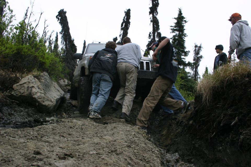 Several people pushing a Jeep Wrangler stuck on a muddy, rocky trail