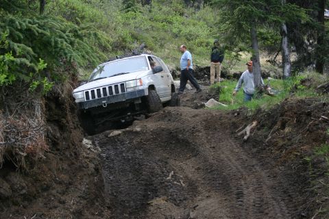 2003 Jeep TJ - A white Jeep Grand Cherokee navigates a challenging muddy trail with several people observing the off-road excursion.