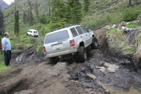 2003 Jeep TJ - A Jeep Grand Cherokee tackles a challenging muddy off-road course, while a white van waits on the trail behind.