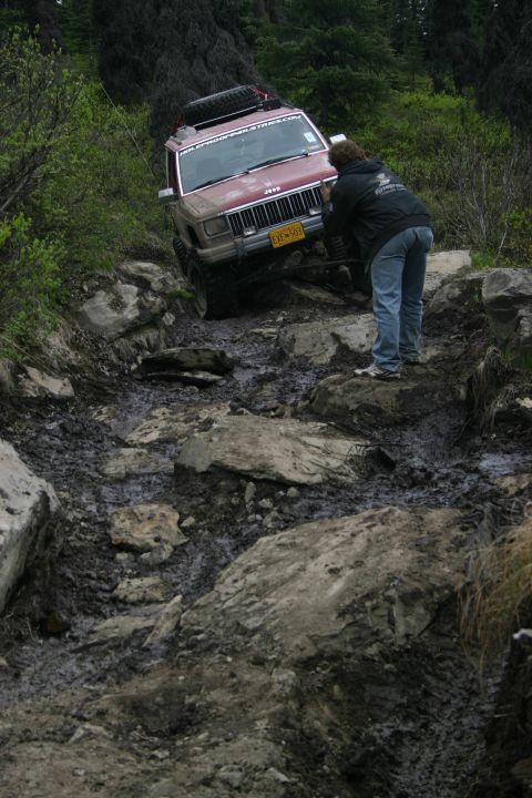 2003 Jeep TJ - A red Jeep Cherokee XJ navigates a challenging rocky and muddy off-road trail, with a person providing guidance.