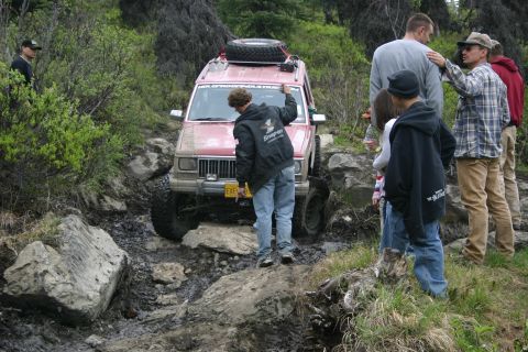 2003 Jeep TJ - A red Jeep Cherokee XJ is shown off-roading on a challenging, muddy, and rocky trail.