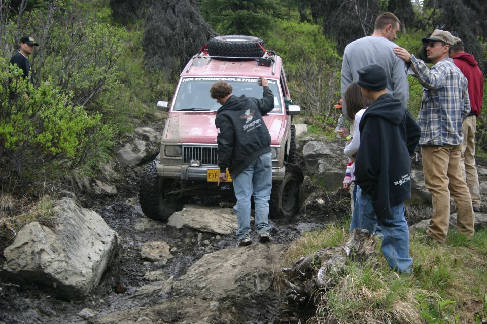Red Jeep Cherokee XJ with large tires navigating a muddy, rocky trail. People stand nearby