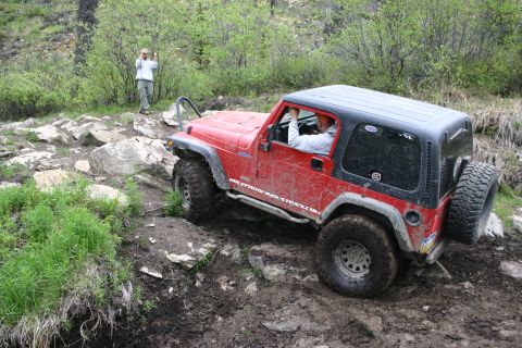 2003 Jeep TJ - A red Jeep TJ is shown off-roading on a challenging, uneven terrain with lush green foliage and large rocks.
