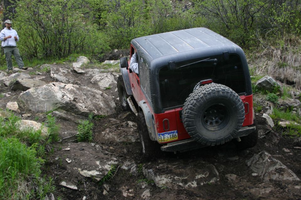 Red Jeep Wrangler TJ navigating a rocky, muddy trail with a person standing on the left