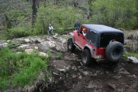 2003 Jeep TJ - A red Jeep Wrangler navigates a rocky off-road trail with assistance from two individuals holding a tow strap.