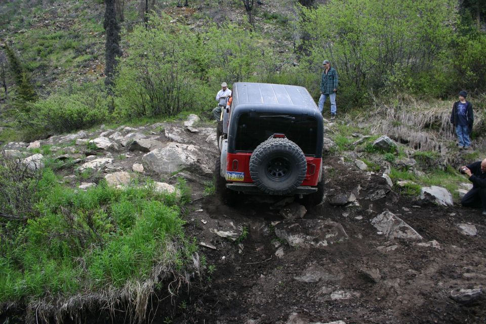 Red Jeep Wrangler navigating a rocky, muddy trail with people watching