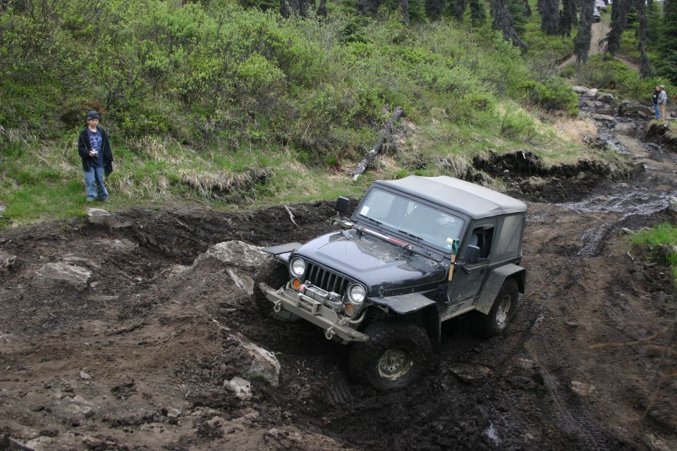 Black 2003 Jeep TJ on a muddy, rocky off-road trail with trees and a person standing on the left