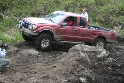 2003 Jeep TJ - A red 2003 Toyota Tacoma navigates a challenging rocky and muddy off-road trail, with a person observing and another person in the driver's…