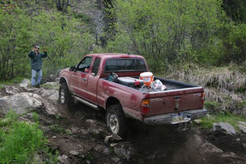 2003 Jeep TJ - A red Toyota Tacoma pickup truck drives off-road on a rocky path, with a person standing nearby.