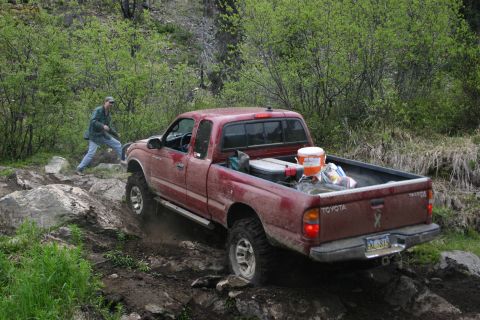 2003 Jeep TJ - A red Toyota Tacoma navigates a challenging rocky and muddy off-road trail with a person walking nearby.