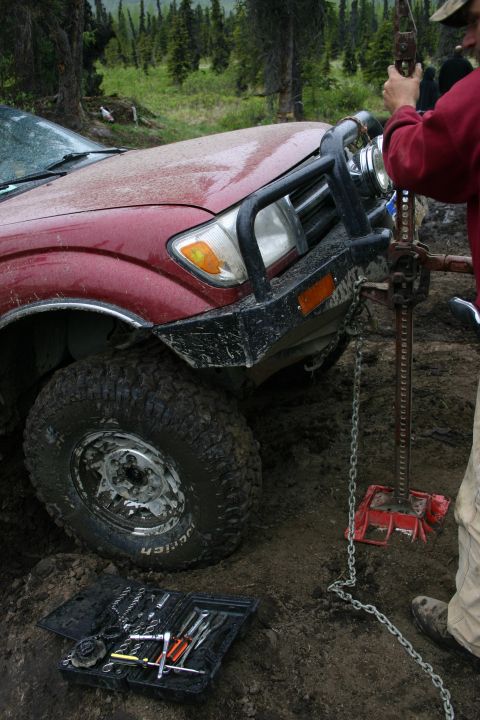 2003 Jeep TJ - A red SUV is stuck in the mud, with a person operating a high-lift jack to free it.