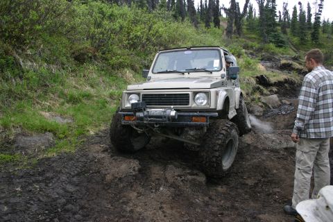 2003 Jeep TJ - A Suzuki Samurai navigates a challenging, muddy off-road course with a person observing the action.