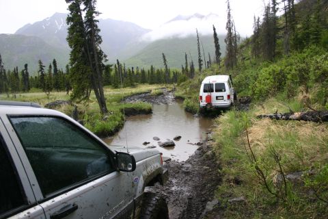 2003 Jeep TJ - Two off-road vehicles traverse a challenging muddy track with water obstacles, surrounded by dense forest and misty mountains.
