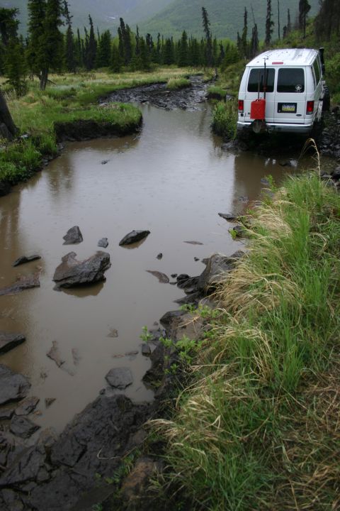 2003 Jeep TJ - A white van navigates a muddy, water-filled trail surrounded by trees and grass.