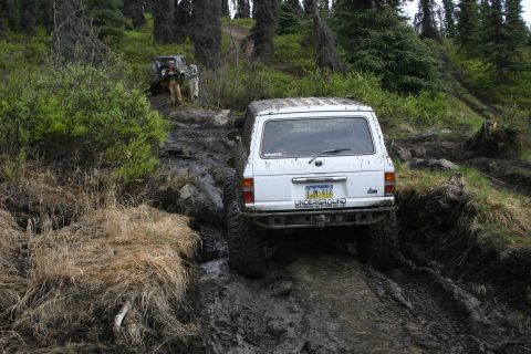 2003 Jeep TJ - A white SUV tackles a challenging muddy path during an off-road event, with another vehicle visible in the background.