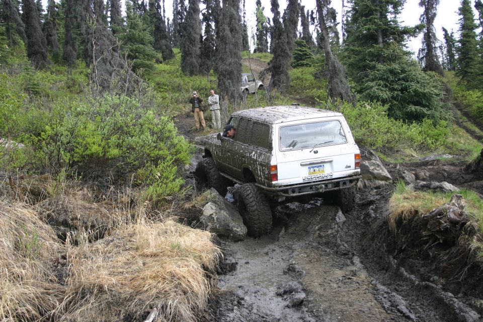 White SUV with large tires on a muddy, rocky trail; another white SUV is visible in the background
