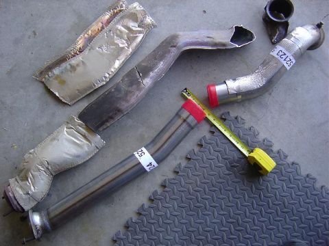 1986 Ford Bronco II - Various exhaust pipes and heat shields are laid out on a textured mat, with a yellow measuring tape nearby.