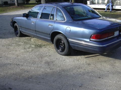 1996 Ford Crown Victoria - A blue sedan is viewed from its rear three-quarter angle, parked on a gravel surface.