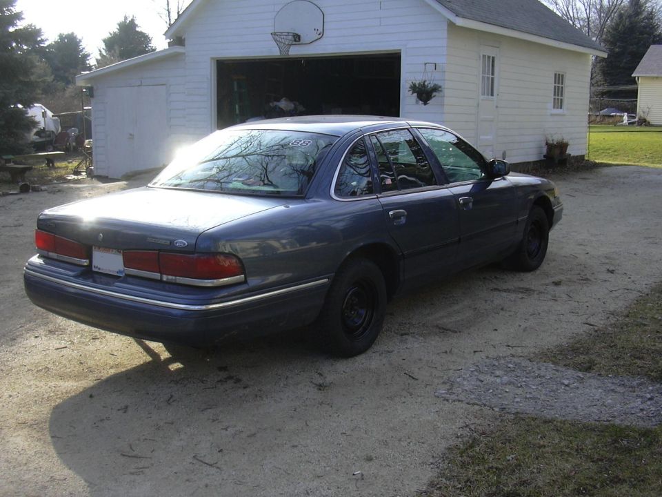 Blue Ford Crown Victoria rear three-quarter view on gravel drive by a white garage, steel wheels