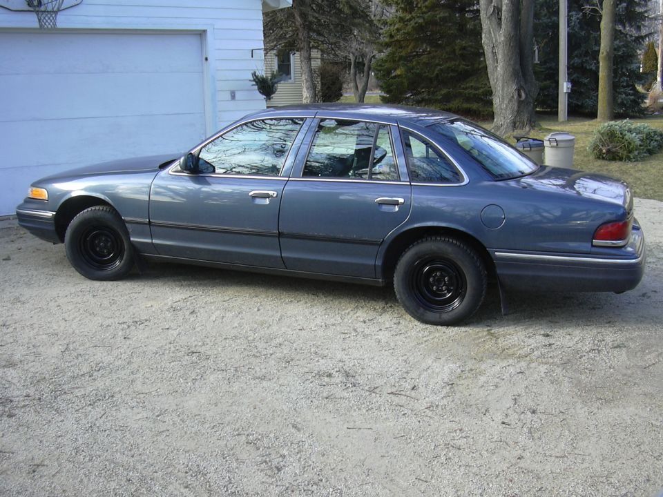 Blue-gray Ford Crown Victoria sedan side view on gravel driveway with black steel wheels