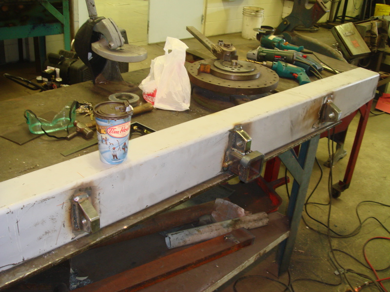 White metal bumper part with visible welds and two hitch receiver mounts, on a workbench with tools and a Tim Hortons cup