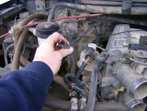 1990 Ford Bronco - Mechanic's hand disconnects a sensor in a dirty engine bay, possibly from a Ford Bronco.